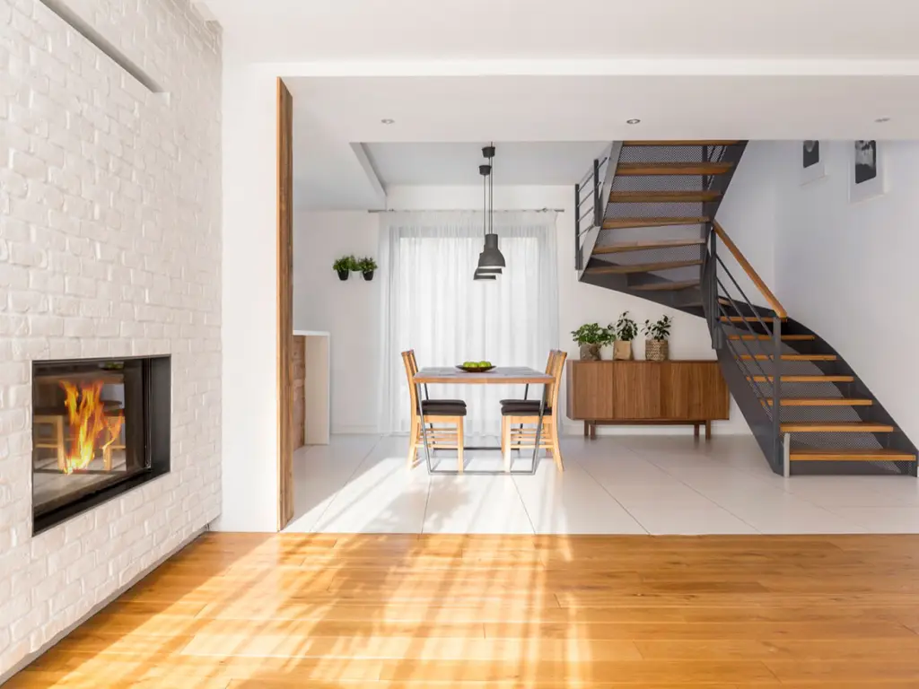 A modern, open-concept living room with a fireplace, dining table, and a staircase leading to an upper floor. The room features white walls, hardwood floors, and large windows that let in natural light.