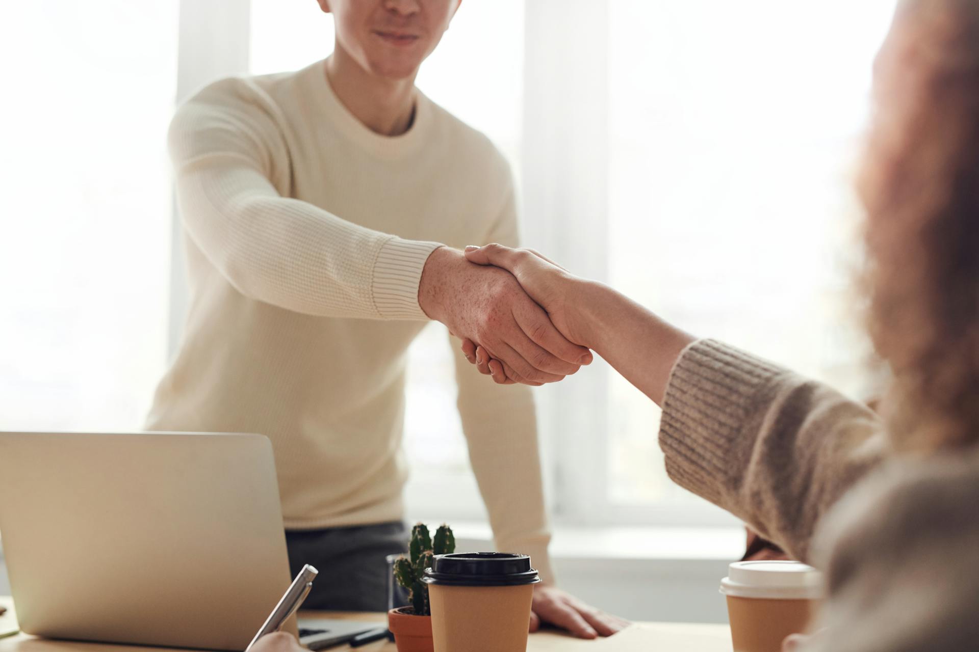 Two people shaking hands over a desk with a laptop on it.