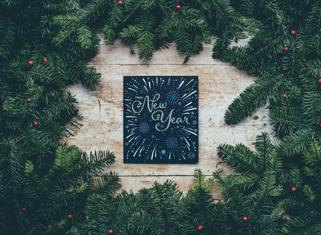 A New Year greeting card featuring glittery text and a fireworks illustration, framed by lush evergreen branches adorned with small red berries.