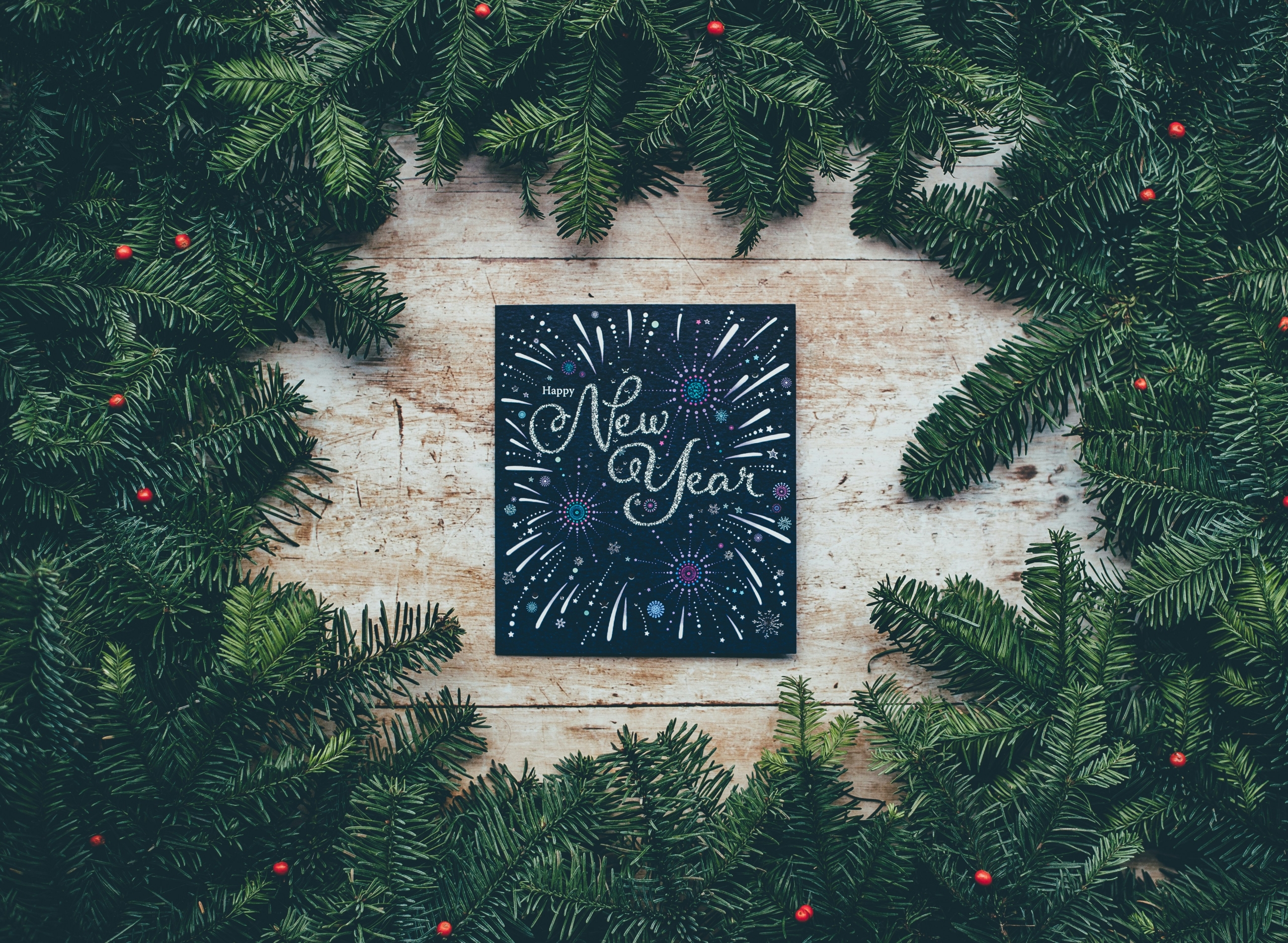 A New Year greeting card featuring glittery text and a fireworks illustration, framed by lush evergreen branches adorned with small red berries.