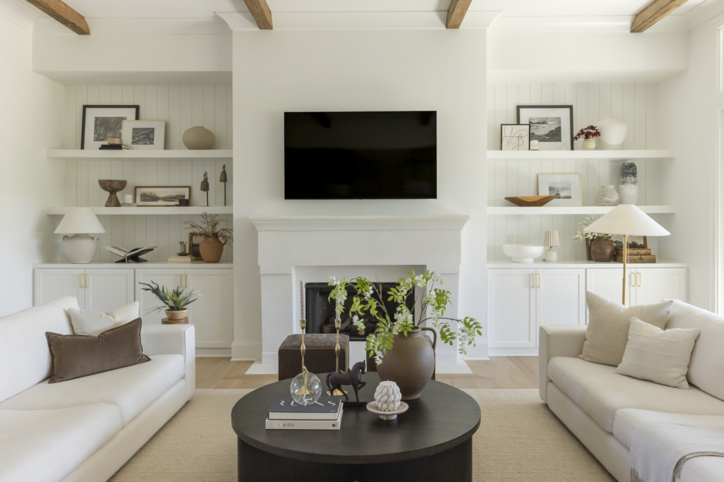 A modern living room with neutral tones, featuring a black round coffee table holding decorative items, two white sofas with beige cushions, a central white fireplace with a mounted flat-screen TV above, and built-in shelves on either side displaying books, framed artwork, plants, and vases.