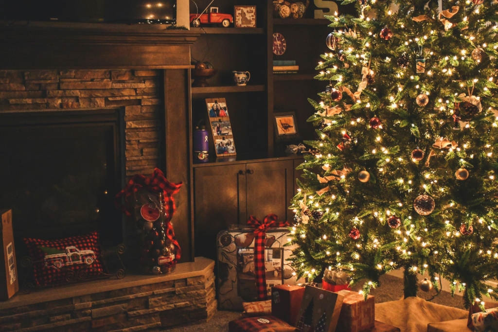 A warmly lit Christmas tree adorned with ornaments stands beside a cozy fireplace and shelves, surrounded by wrapped gifts.