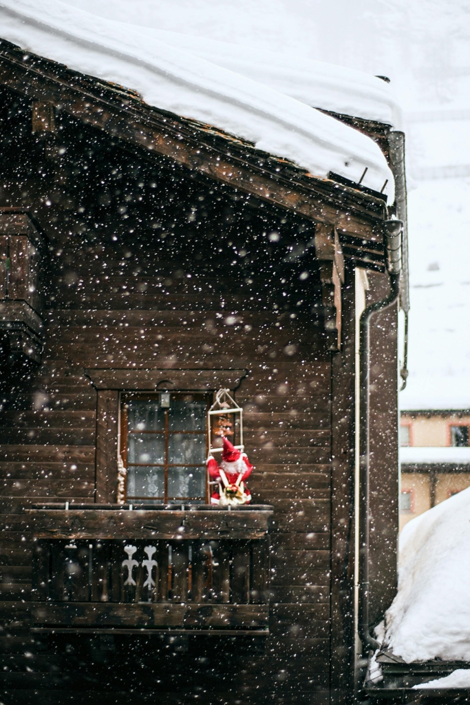 A wooden chalet balcony covered in snow, with a Santa Claus decoration sitting in a lantern, surrounded by gently falling snowflakes.