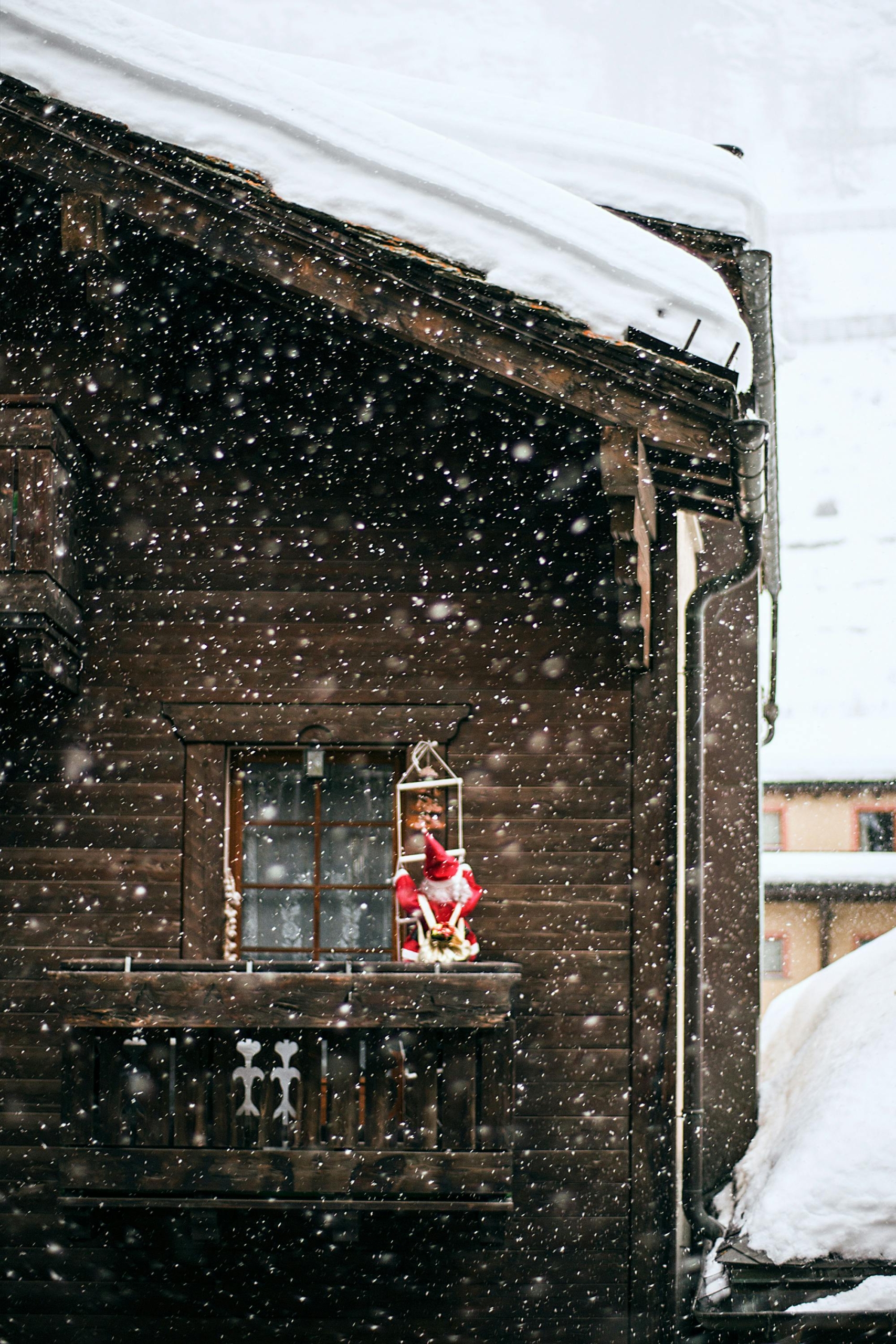 A wooden chalet balcony covered in snow, with a Santa Claus decoration sitting in a lantern, surrounded by gently falling snowflakes.