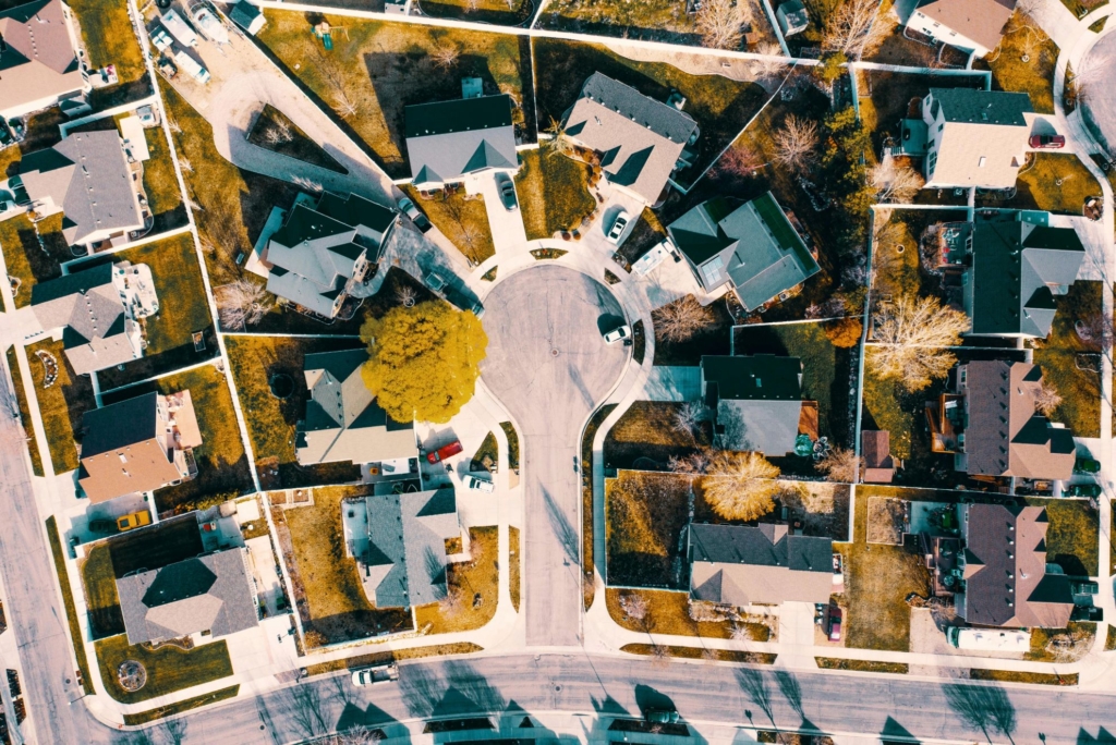 Aerial view of a suburban neighborhood featuring a cul-de-sac surrounded by houses with driveways, lawns, and scattered parked cars. The warm sunlight casts long shadows across the streets and rooftops.