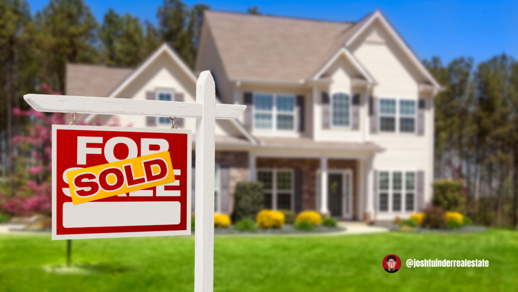 A red and white "For Sale" sign with a large yellow "SOLD" sticker is displayed in the foreground, indicating a recently sold home. Behind it, a large, two-story suburban house with beige siding, multiple windows, and manicured landscaping is slightly blurred. The background features tall trees and a clear blue sky. In the bottom right corner, a logo and the handle "@joshtuinderrealestate" are visible.