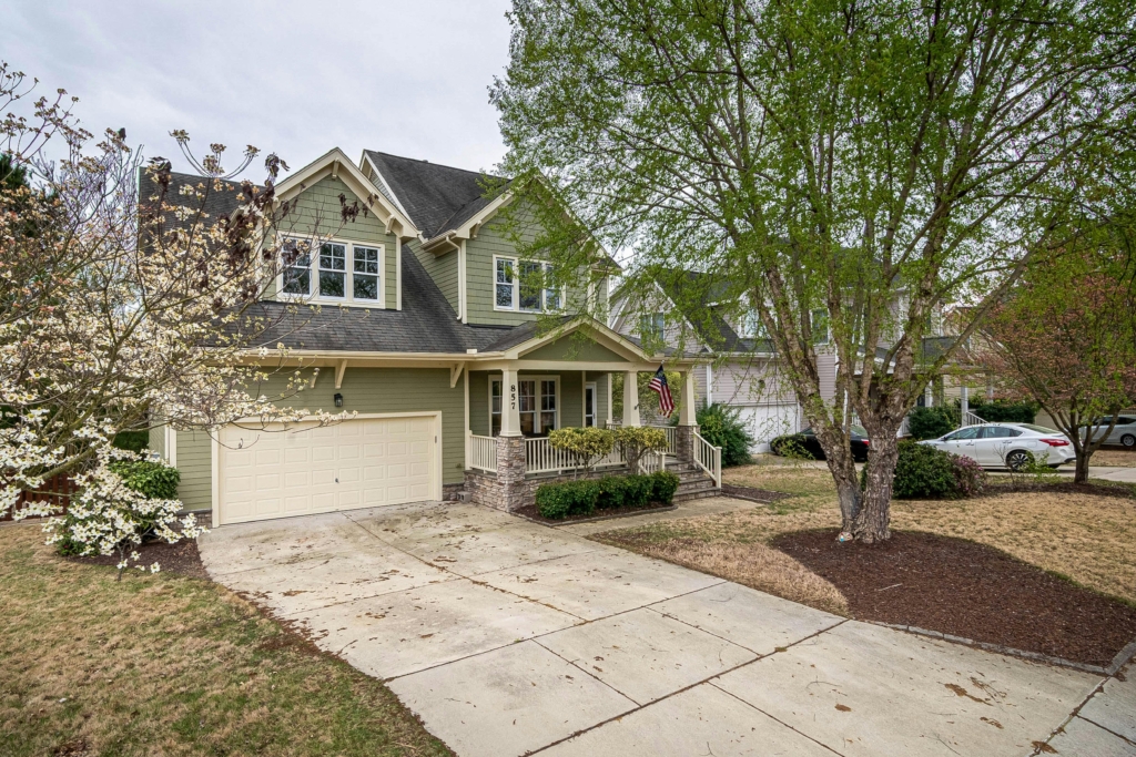 A two-story suburban home with sage green siding, white trim, and a covered front porch. The house has a two-car garage, a concrete driveway, and a well-maintained front yard with trees and flowering plants. An American flag hangs from the porch railing. Neighboring houses and parked cars are visible in the background.