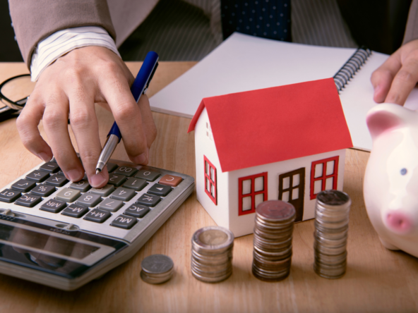 A close-up of a person in business attire using a calculator at a desk, with a small model house, stacked coins, a piggy bank, and a notebook in view, symbolizing financial planning, mortgage calculations, or real estate investment. Symbolizing budgeting during a housing recession.