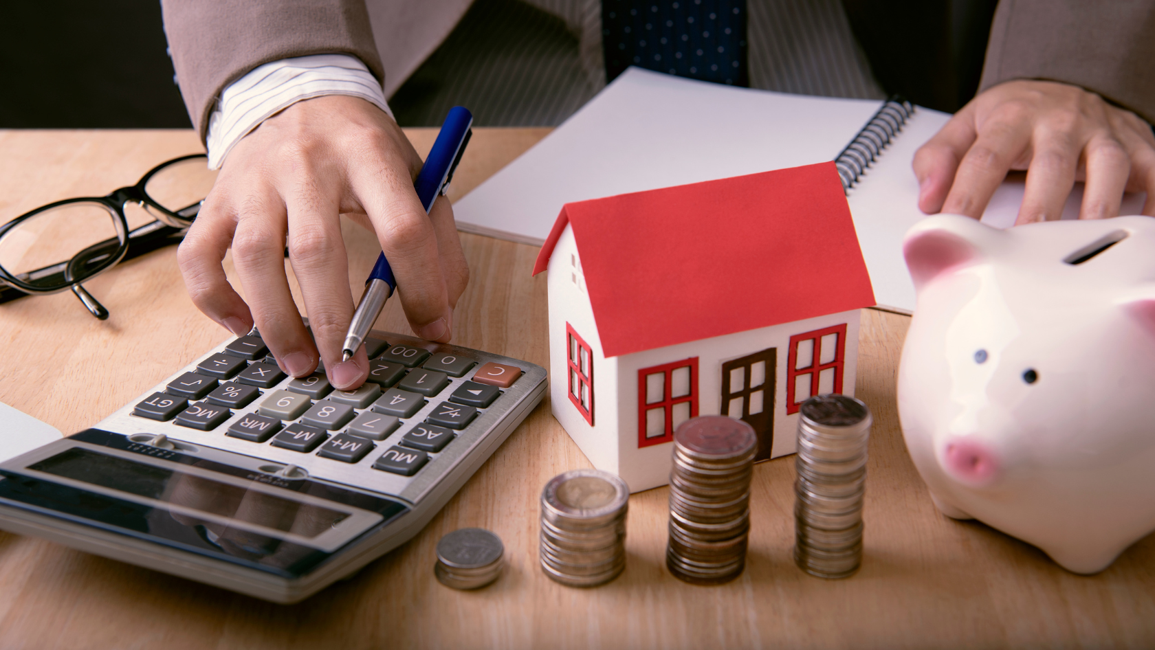 A close-up of a person in business attire using a calculator at a desk, with a small model house, stacked coins, a piggy bank, and a notebook in view, symbolizing financial planning, mortgage calculations, or real estate investment. Symbolizing budgeting during a housing recession.