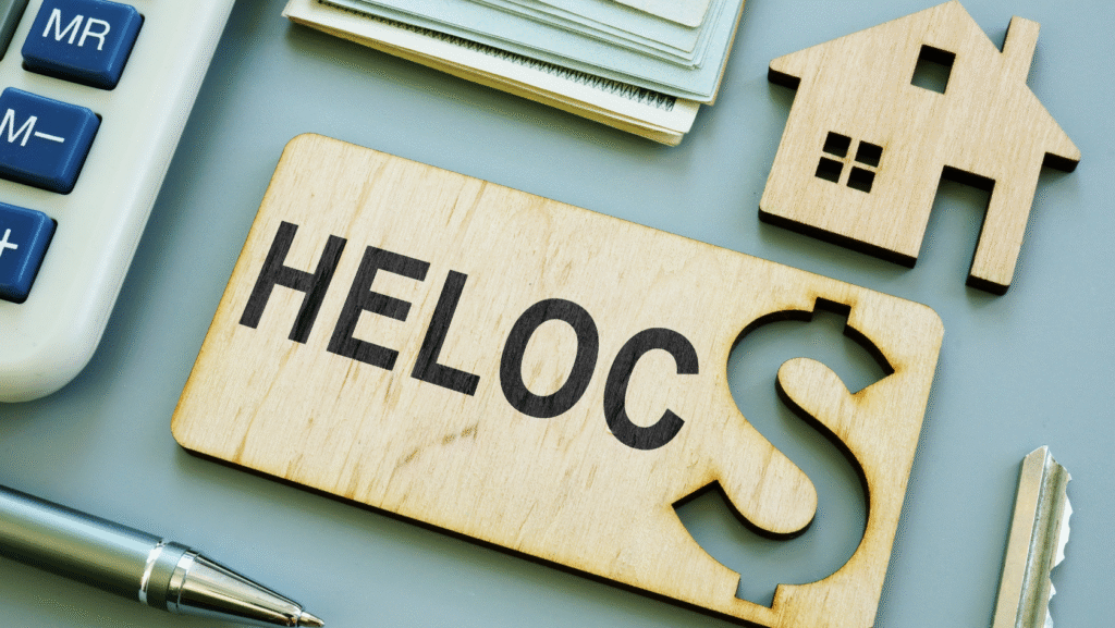 Wooden block with the word "HELOC" and a dollar sign cutout, surrounded by a house-shaped cutout, a stack of money, a calculator, a pen, and a key.