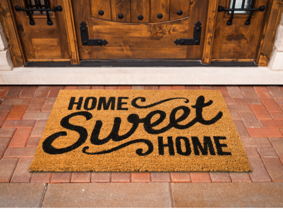A doormat with the phrase “Home Sweet Home” in bold black letters sits in front of a wooden front door on a red brick porch.