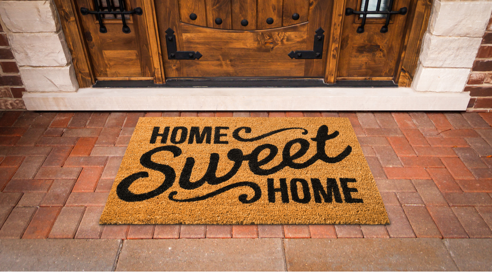 A doormat with the phrase “Home Sweet Home” in bold black letters sits in front of a wooden front door on a red brick porch.