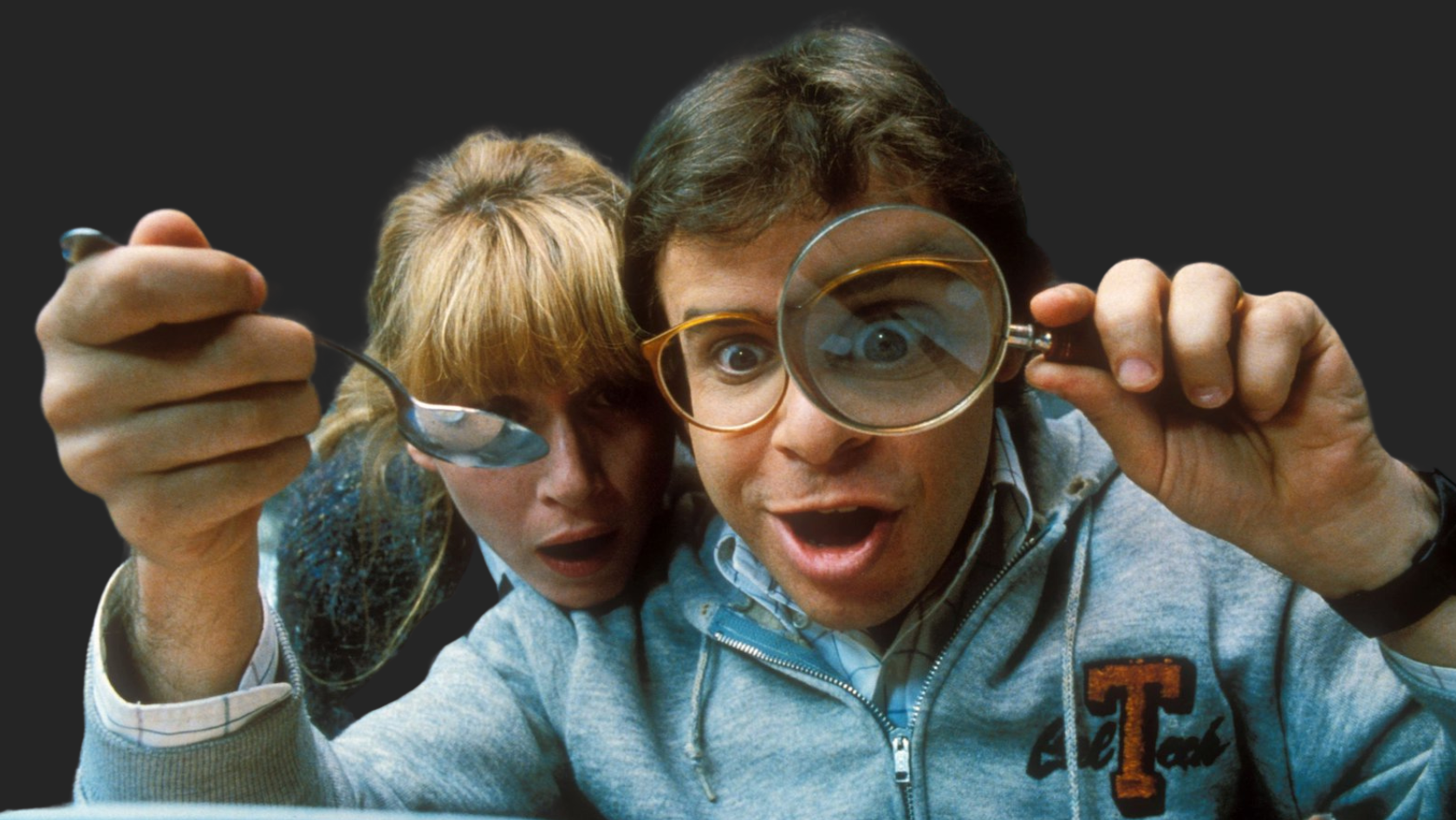 A man and woman peer closely at something, the man holding a magnifying glass and the woman a spoon, both with exaggeratedly amazed expressions.