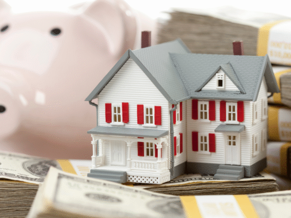 A miniature model of a white house with red shutters sits atop stacks of U.S. dollar bills, with a pink piggy bank blurred in the background.