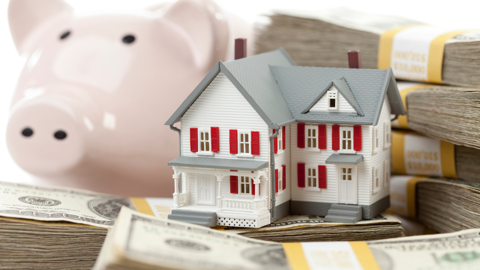 A miniature model of a white house with red shutters sits atop stacks of U.S. dollar bills, with a pink piggy bank blurred in the background.